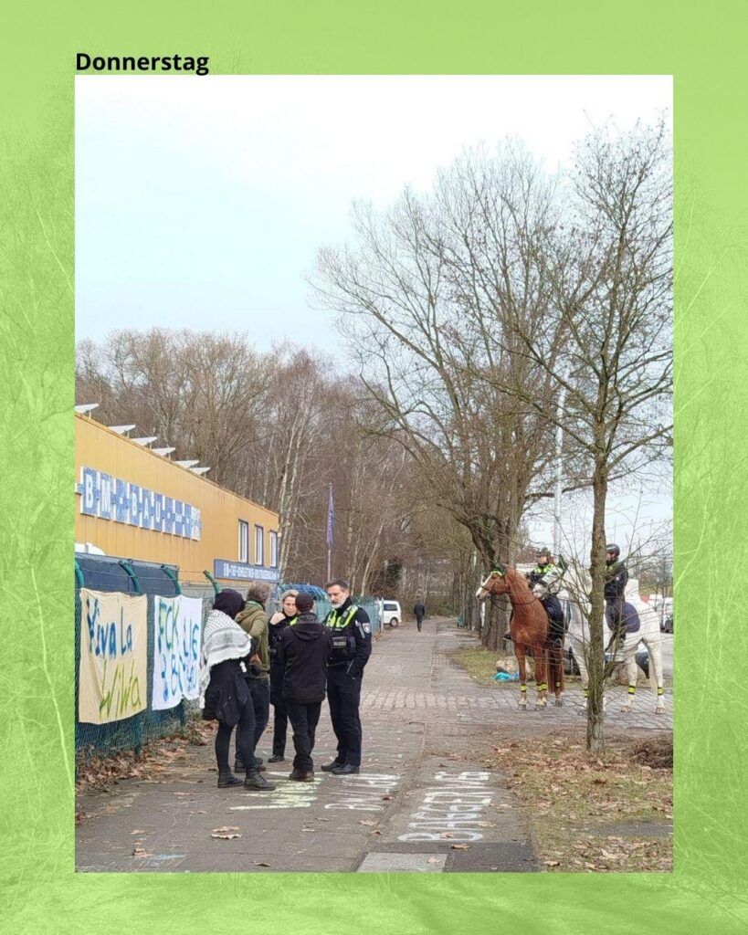 Foto vom Eingangstor von BM-Bau. Links stehen drei Aktivistis und zwei Cops und diskutieren, rechts stehen zwei Polizeipferde und warten. 
